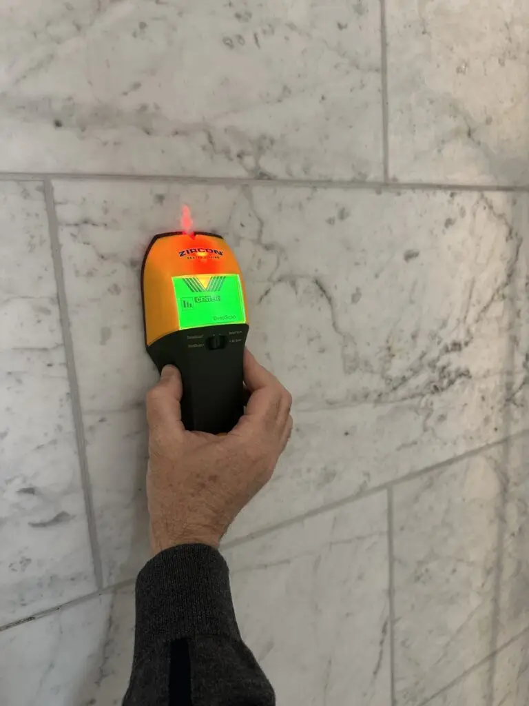 Stud finder scanning a marble bathroom wall to locate safe mounting points for grab bar installation.